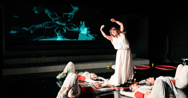 Schauspielerin Juliana Götze tanzt mit erhobenen Armen in einem roten Ring. Mit den Händen wird dieser gehalten von 4 Chor-Darstellerinnen, die auf dem Rücken am Boden liegen. Eine Live-Kamera filmt sie von oben. Das Bild wird auf die Rückwand projiziert.