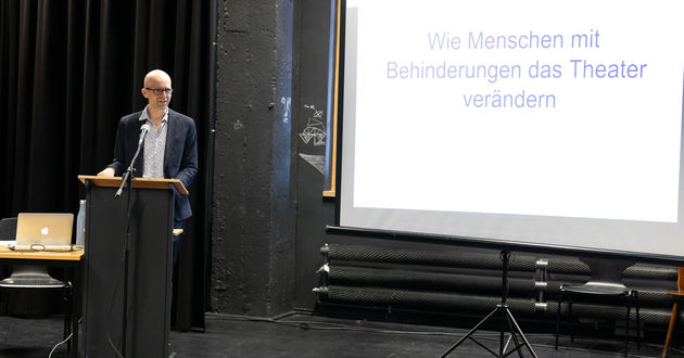 Georg Kasch steht an einem Redner*innenpult und beginnt  einen Vortrag. Rechts neben ihm steht eine große Leinwand. Darauf ist  der Titel seines Vortrags projiziert: „Wie Menschen mit Behinderungen das Theater verändern“.