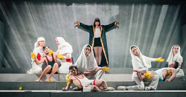 Auf einer Tribüne bilden 8 Schauspieler*innen ein Standbild einer Pietat. Im Hintergrund breitet Schauspielerin Hieu Pham ihre Arme aus, wie Jesus am Kreuz.