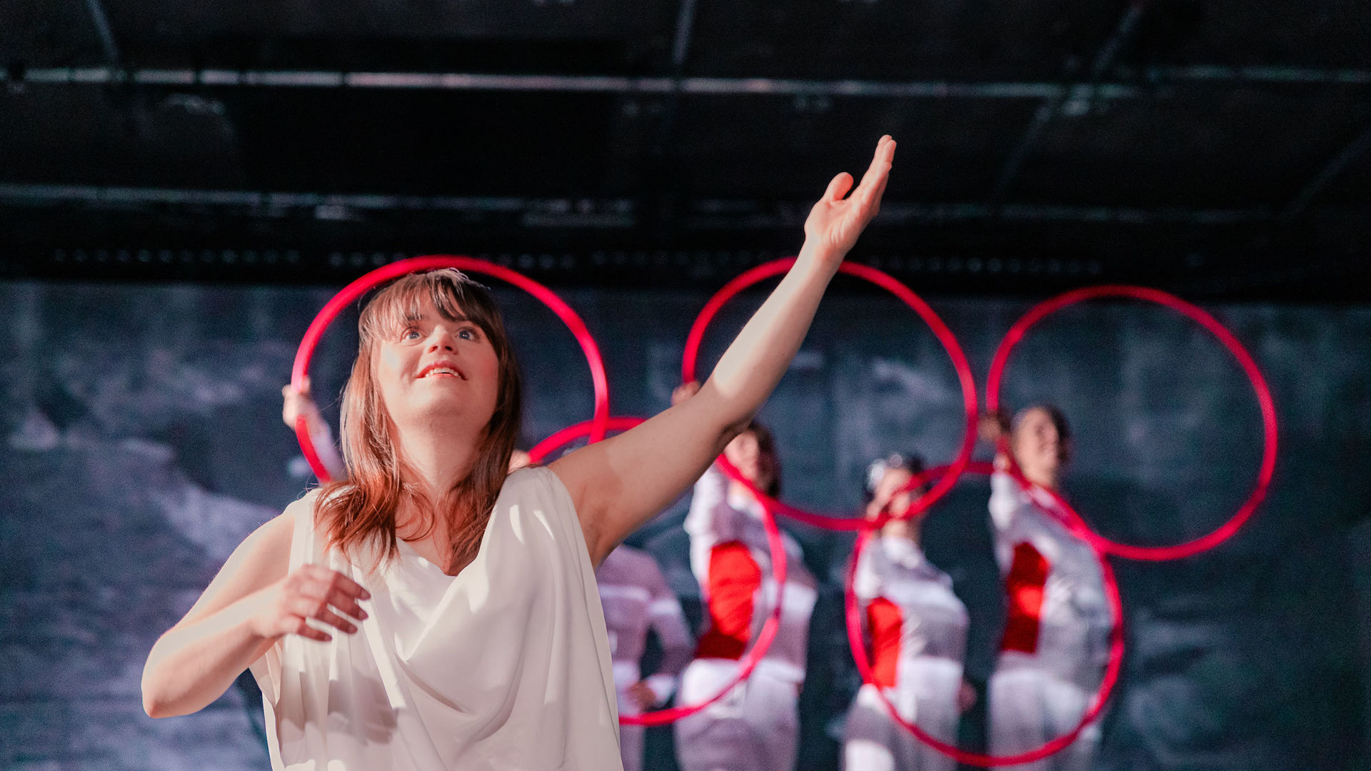 Schauspielerin Juliana Götze hebt ihren linken Arm und schaut lächelnd zur Decke. Im Hintergrund formen die 5 Chor-Darstellerinnen mit 5 roten Ringen das Emblem der Olympischen Spiele.