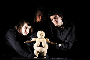Drei, in schwarz gekleidete Puppenspielerinnnen, hinter einer beleuteten Puppe auf einem Tisch, die der "Venus von Willendorf" Stautuette ähnelt