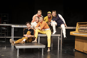 Sechs Schauspierler*innen, in organge-weiß-gelb-schwarz gehaltenen Kostümen, die in unterschiedlichen Körperhaltungen auf Bühnenpodesten positioniert sind