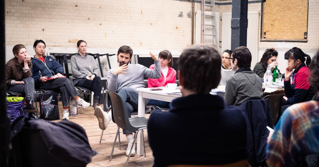 Offene Leseprobe der „Sportstück“-Inszenierung am RambaZamba Theater. Die Ensemblemitglieder  sitzen an einem Tisch in der Mitte der Probebühne des RambaZamba-Theaters. Jacob Höhne sitzt an der Ecke des Tisches, spricht und gestikuliert.