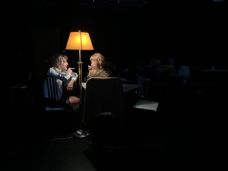 Zwei junge Frauen, nur von einer Stehlampe beleuchtet, sitzen an einem Tisch, rauchend und lachend. 