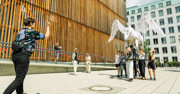 Draußen vor der HfS Ernst Busch führen 7 Spieler*innen eine große weiße Pegasus-Puppe. Mit Stöcken führen sie die Großpuppe über ihren Köpfen und bewegen sie auf die Gastprofessorin Roscha Säidow zu, die die Puppenführung anleitet.