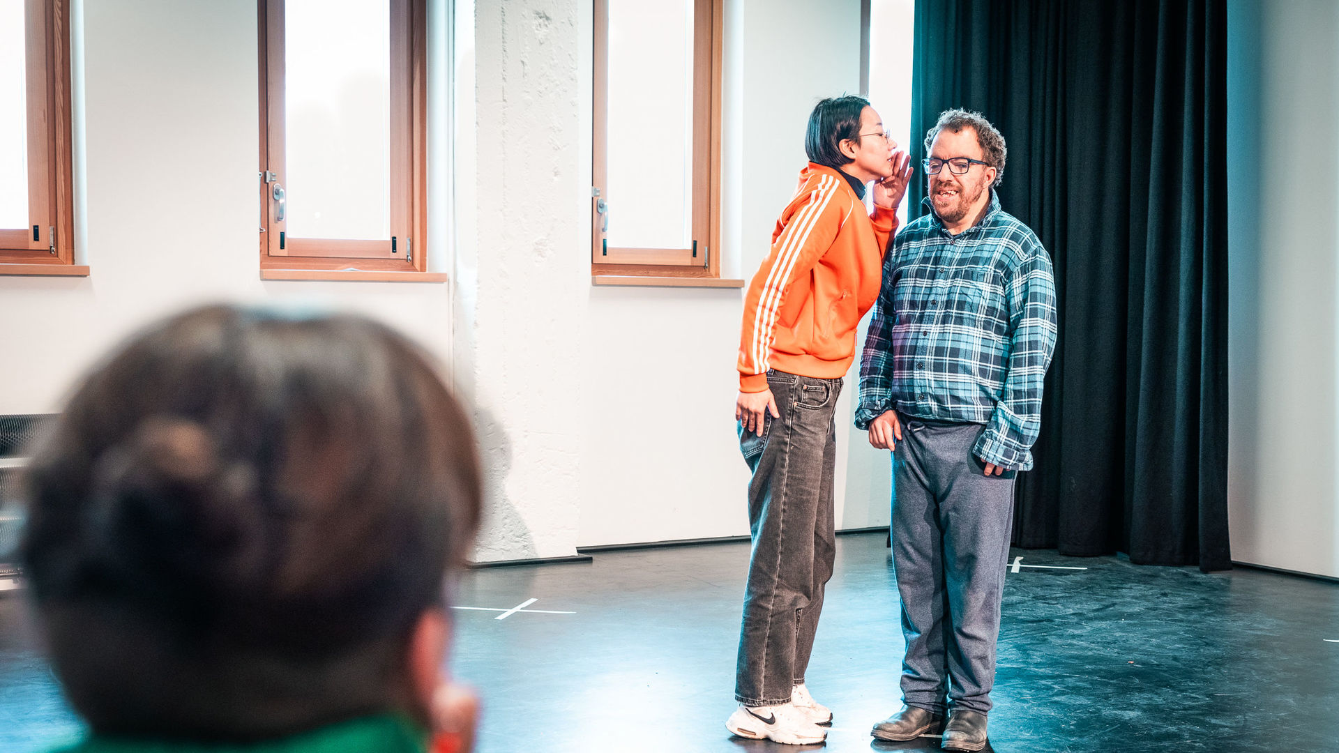 Workshop mit Nele Jahnke zum Impuls I: Regiearbeit mit einem mixed abled Ensemble Zwei Workshopteilnehmer*innen improvisieren eine Szene. Eine Person flüstert der anderen etwas ins Ohr.