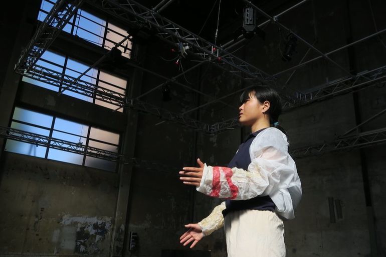 Foto einer Tänzerin, stehend mit angewinkelnden Armen, in weiß-schwarzen Kostüm mit roten Musern an den Ärmeln. Perspektive von unten, sodass viel Raum zu sehen ist, eine Halle mit Bühnentechnik und großen Fenstern 