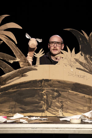Eine Bühnenbild aus Pappe, dass eine Insel mit Palmen darstellt. Dazwischen der Kopf eines Puppenspielers mit schwarzer Hornbrille, der eine Sockenpuppe hochält, im Stile eines Schweins