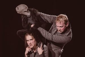 Eine Puppe mit Echsenkopf und Menschenköper, die auf den Schultern einer Puppenspielerin hockt, während sie von einem weiteren Pupenspieler, daneben stehend, gesteuert wird. Alle drei Figuren haben dunkle, graue Kostüme an 