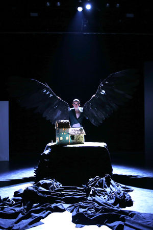 Puppenspielerin mit großen schwarzen Flügeln, in düsterem Licht vor dem Modell einer Mühle. Davor ein schwarzer Fluss, der mittels eines schwazen Tuches dargestellt wird. Schwarze Federn auf dem Boden 