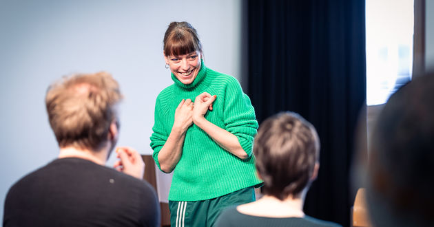 Nele Jahnke steht lächelnd vor den Workshopteilnehmer*innen, die ihr gegenüber auf Stühlen sitzen.