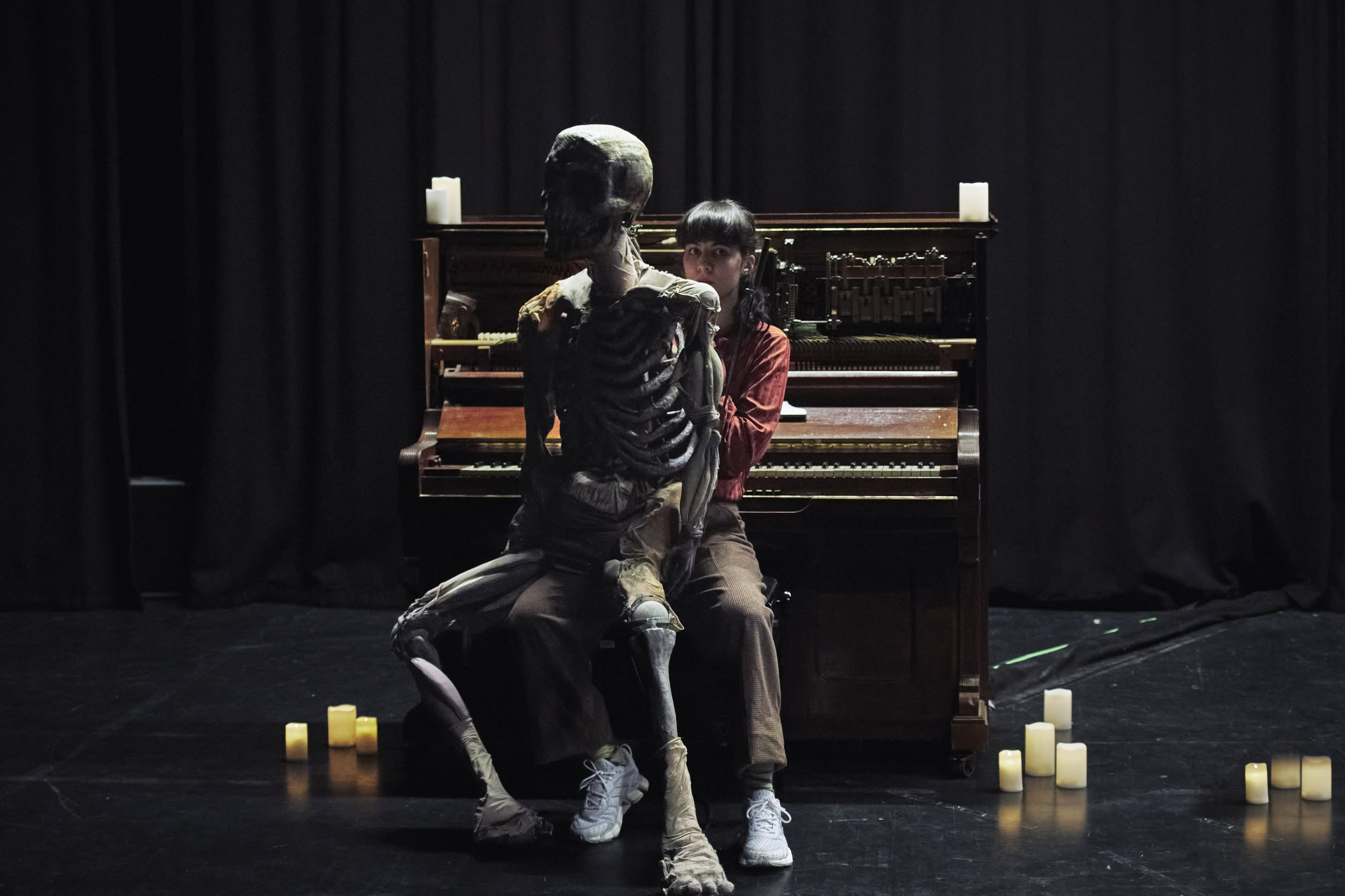Große Skelettpuppe mit Puppenspielerin vor Klavier auf dunkler Bühne 