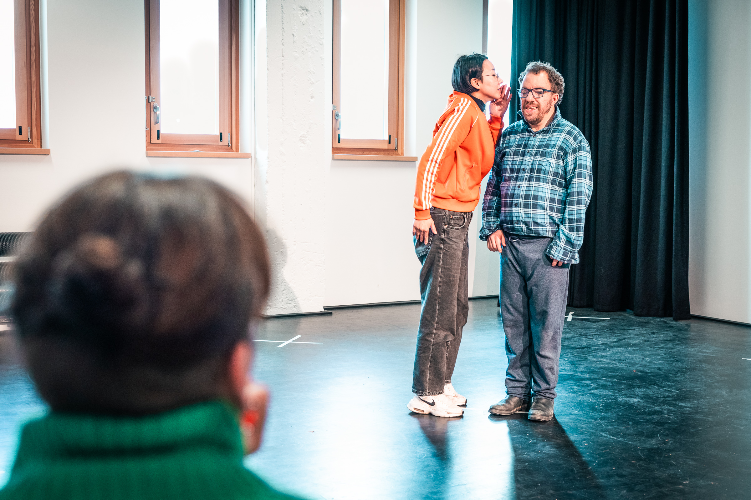 Zwei Workshopteilnehmer*innen improvisieren eine Szene. Eine Person flüstert der anderen etwas ins Ohr. 