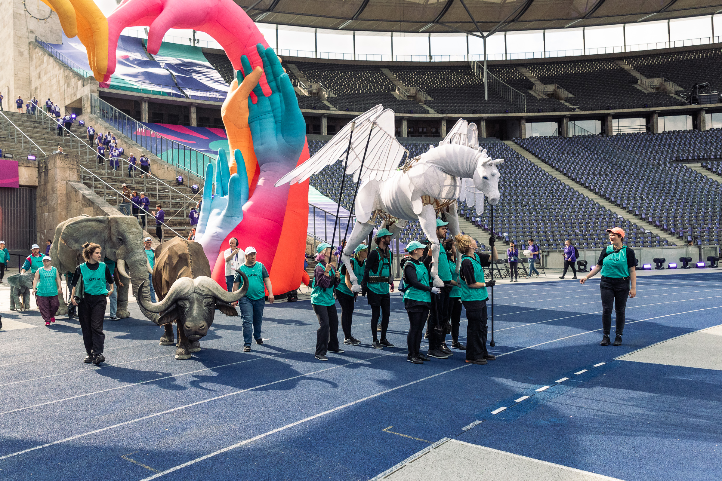 Die Spielenden des Walking Acts führen vier Großpuppen Pegasus, Büffel und zwei Elefanten über die Rennbahn in das Olympia-Stadion. Gastprofessorin Roscha Säidow steht vorne und leitet sie an.